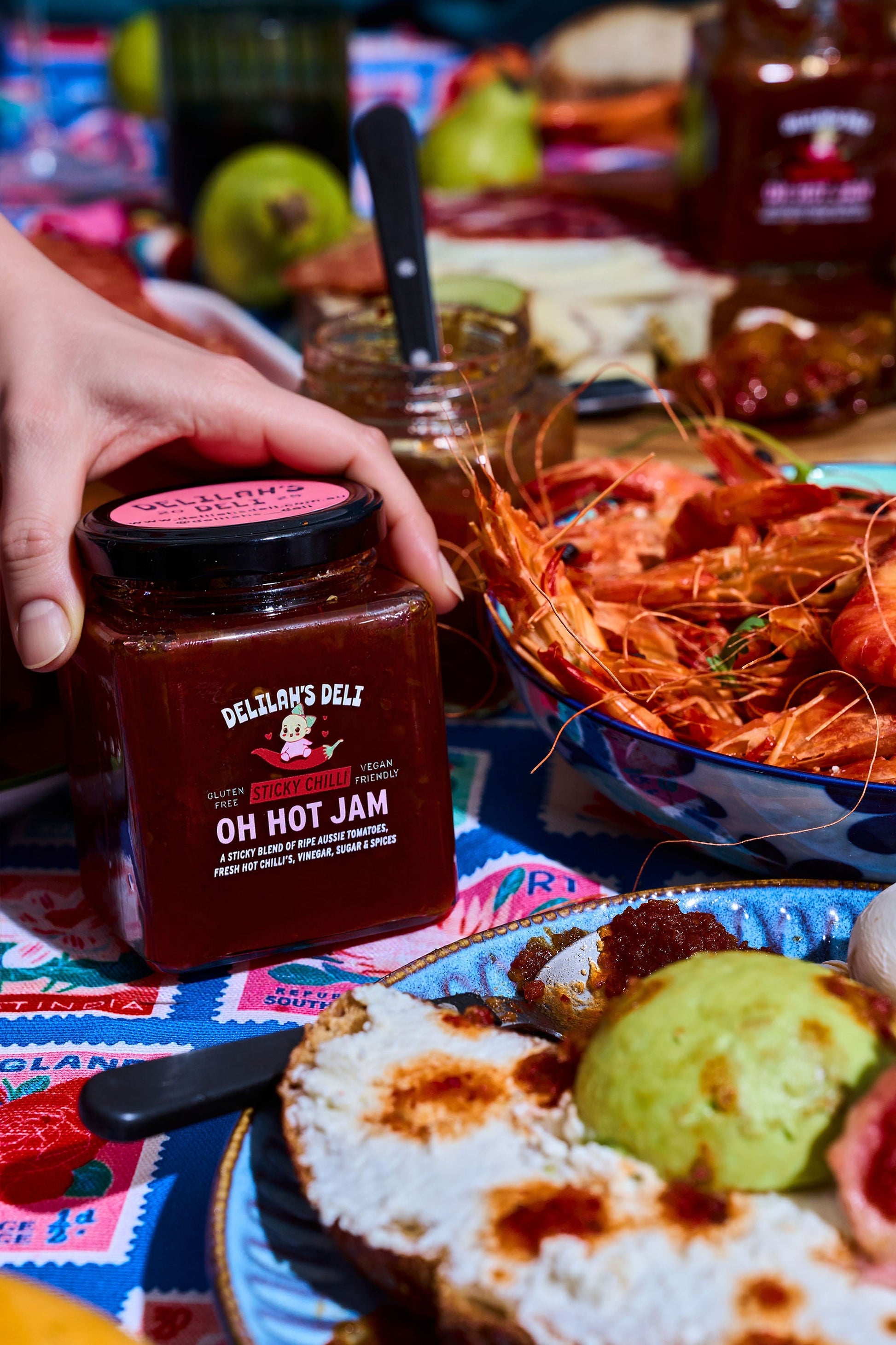 Person holding a jar of Del Monte Deli Oh Hot Jam with a table of food in the background