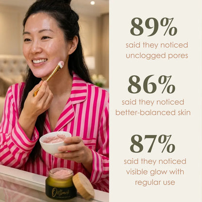 A smiling woman in pink striped pajamas applies a pink skincare cream from a small bowl with a gold-handled brush while standing in front of a mirror. A jar of the product sits on the counter below.